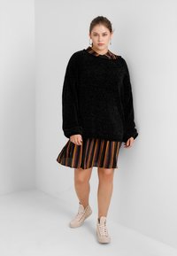 Black, textured sweatshirt layered over a multi-colored striped dress with pleats, paired with light-colored high-top sneakers.