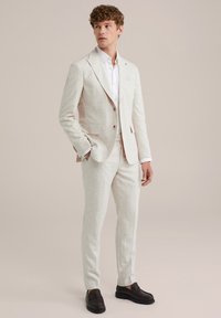 Világos bézs vászon öltöny, slim-fit kialakítással, egysoros zakóval, kihajtható gallérral és illő nadrággal. Fehér inggel és barna mokaszinokkal párosítva.