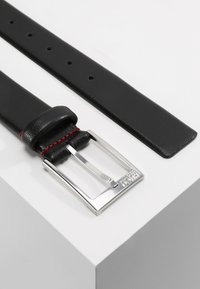 Cinturón de cuero negro con costuras rojas, que presenta una hebilla rectangular de plata y múltiples agujeros de ajuste para el tamaño. Textura suave.