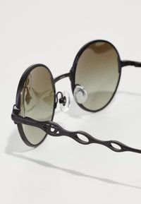 Ochelari de soare rotunzi cu lentile cu umbrire întunecată, ramă din plastic negru și un design unic al curelei, cu decupaje ovale pe templuri.