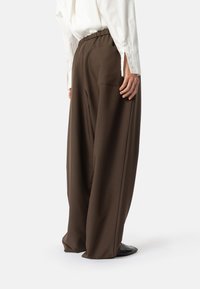 Bruine wijde broek met elastische tailleband, voorzien van een achterzak en een gladde, vloeiende stoftextuur. Gecombineerd met een wit overhemd.