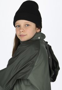 Grøn vandtæt jakke med høj krave og trykknaplukninger, parret med en sort strikket beanie. Glat tekstur og tætsiddende design.