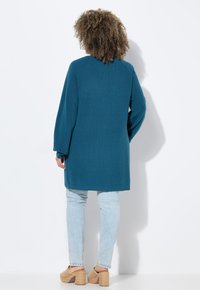 Dlouhý tyrkysový pletený cardigan s texturovaným vzorem, širokými rukávy a bočními rozparky, spárovaný se světlými modrými skinny džíny a béžovými platformovými sandály.