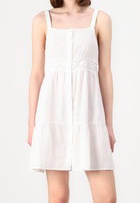 Robe d'été blanche avec bretelles réglables, fermeture à boutons sur le devant et détail en dentelle à la taille. Jupe évasée avec design à étages, fabriquée en tissu léger.