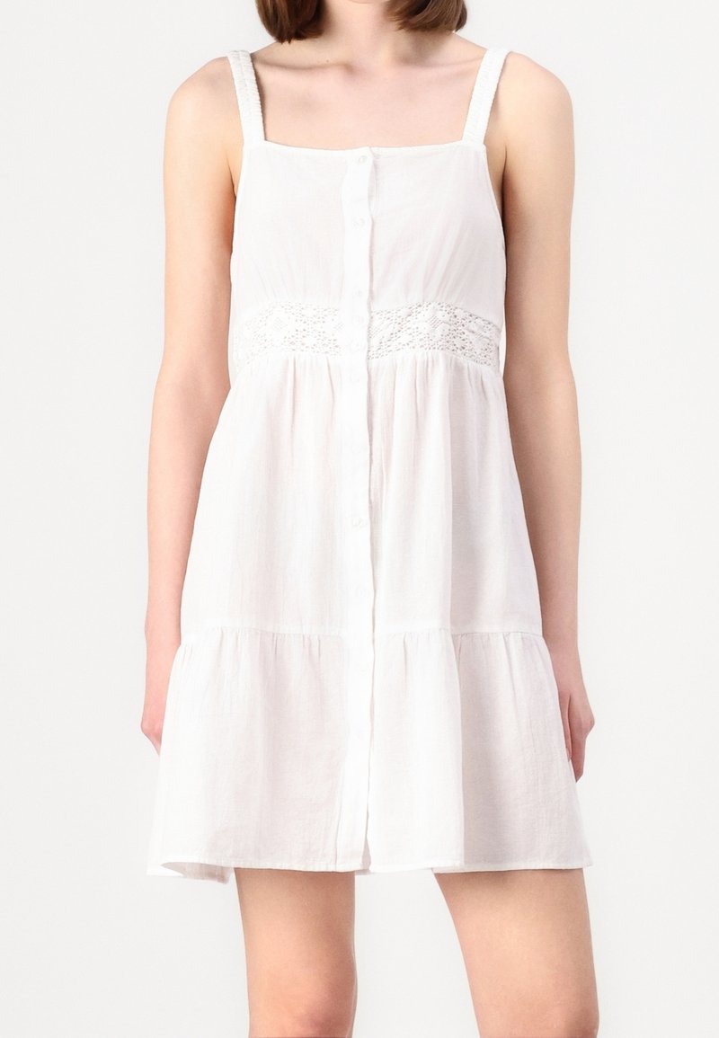 Robe d'été blanche avec bretelles réglables, fermeture à boutons sur le devant et détail en dentelle à la taille. Jupe évasée avec design à étages, fabriquée en tissu léger.