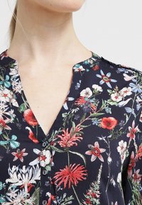 Bluse i marineblå stof med blomsterprint, featuring V-udskæring, knappelukning og farverige blomster i forskellige størrelser og former.