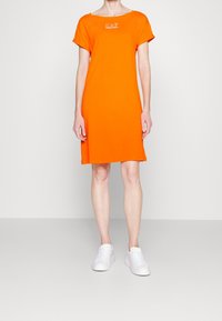 Person klädd i en ljusorange kortärmad klänning med "EA7 Emporio Armani"-logotyp och vita sneakers, stående mot en plain ljus bakgrund.