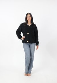 Cardigan nero in pelucchino con grandi bottoni dorati e due tasche frontali, indossato sopra jeans blu. Texture morbida e vestibilità rilassata.