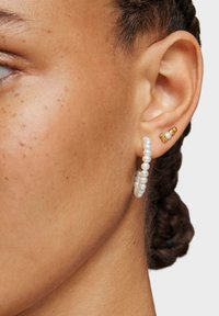 Zbliżenie ucha osoby z zaplecionymi włosami, noszącej białe kolczyki-koła z koralikami oraz trzy małe złote i perłowe kolczyki wkrętki.