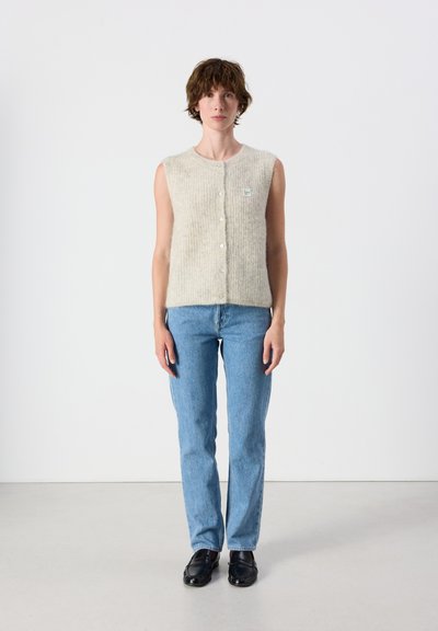 Adulte aux cheveux courts et bruns portant un gilet beige sans manches, un jean bleu et des chaussures noires, debout devant un fond blanc uni.