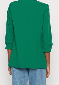 Personne portant un blazer vert vif avec les manches retroussées et un jean bleu clair, vue de dos sur un fond uni.