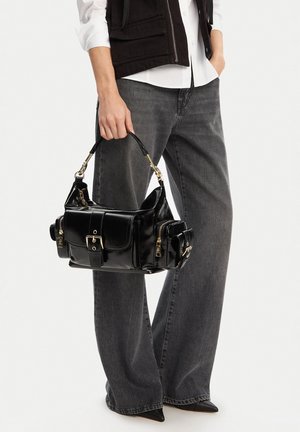 Osoba ubrana w czarną kamizelkę, białą koszulę i szare dżinsy trzyma błyszczącą czarną torebkę z wieloma kieszeniami na suwak i złotymi okuciami.