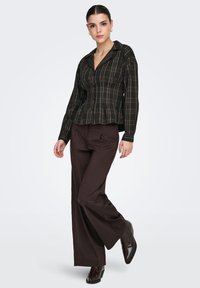 Chemise à manches longues à carreaux noirs et verts avec un col, associée à un pantalon large marron foncé et des bottines noires brillantes.