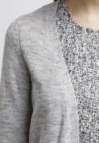 Ljusgrå cardigan gjord av mjukt stickat tyg med subtil textur. Lager över en svartvit strukturerad topp. Enkel design, öppen fram.