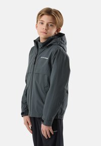 Fant z blond kratkimi lasmi, oblečen v sivega kapucarja z zadrgo in črne hlače, stoji na belem ozadju.