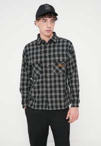 Chemise en flanelle à carreaux noir et blanc avec des boutons dorés, comprenant deux poches avant et un logo orange sur la poitrine gauche.