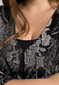 Zwarte kanten cardigan met zilverkleurig bloemenpatroon, met een losse pasvorm en korte mouwen, gedragen over een zwart topje. Textuurdetail zichtbaar.