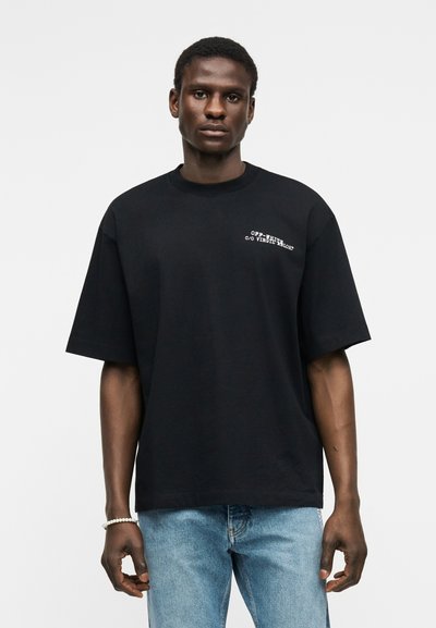 Άνδρας που φοράει μαύρο υπερμεγέθες T-shirt με λευκό κείμενο στο στήθος και ανοιχτόχρωμο τζιν, στέκεται μπροστά σε λευκό φόντο.