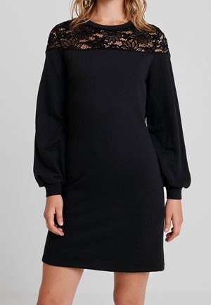 Vrouw die een zwarte jurk met lange mouwen draagt, met een bloemrijke kanten paneel over de borst en schouders, staand tegen een effen achtergrond.