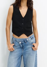 Gilet noir sans manches avec un décolleté en V profond, cinq boutons sur le devant et deux poches latérales, associé à un jean ample bleu clair.