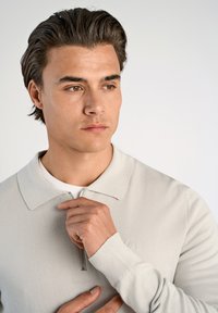 Maglione grigio chiaro con zip e una consistenza morbida, dotato di colletto e polsini a costine. Il modello tiene la zip, indossando una camicia bianca sotto.