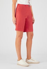 Pantalones cortos ajustados rojos con una textura suave, de longitud hasta el muslo y cierre frontal con botón. Combinados con zapatillas blancas con acentos dorados.