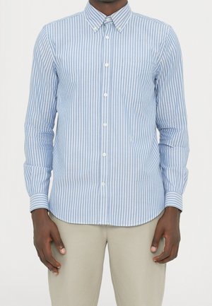 Homme portant une chemise à boutons rayée verticalement bleu clair et blanc avec un pantalon beige, debout les bras détendus le long du corps.