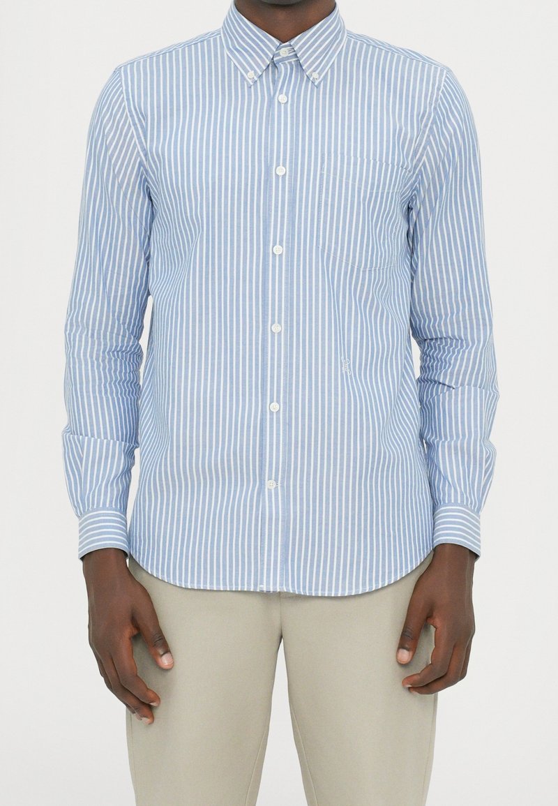 Homme portant une chemise à boutons rayée verticalement bleu clair et blanc avec un pantalon beige, debout les bras détendus le long du corps.