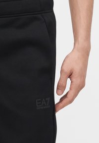 Δεξί χέρι δίπλα σε μαύρο παντελόνι με διακριτικό λογότυπο EA7 Emporio Armani κοντά στην τσέπη, με απλό ανοιχτόχρωμο φόντο.