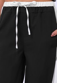 Schwarze Jogginghosen mit elastischem Bund, weißem Kordelzug, seitlichen Taschen und kontrastierenden weißen Seitenstreifen. Glatte Textur.