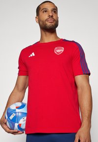 T-shirt rouge Adidas avec des rayures bleues sur les épaules, affichant un logo d'Arsenal. Le mannequin tient un ballon de football aux motifs bleu et blanc.