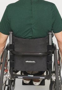 Černý vozík pro invalidy s označenou oporou na záda, stříbrnými koly a opěrkami na ruce. Osoba v zeleném tričku a bílých botách s hnědými detaily.