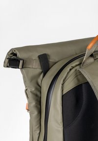 Gros plan sur un coin d'un sac à dos vert à enroulement avec une fermeture éclair noire, une sangle, une boucle et un petit détail de poignée en cuir marron.