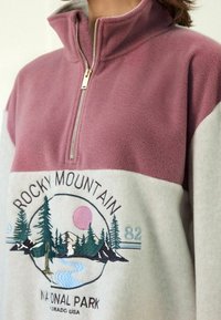 Bluza polarowa z półzamkiem, z różową górną i kremową dolną częścią. Haftowany wzór drzew i gór, z złotym zamkiem.