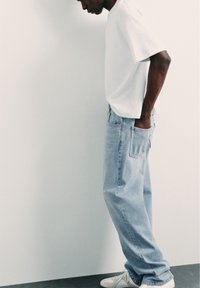 Homme portant un t-shirt blanc, un jean bleu clair et des baskets blanches, debout de côté, les mains dans les poches arrière, contre un mur uni.