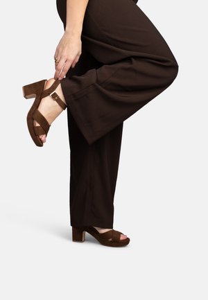 Persona che regola sandali marroni in camoscio con tacco a blocco indossando pantaloni marroni a gamba larga coordinati su uno sfondo semplice.