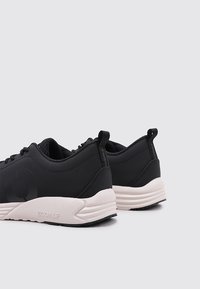 Las zapatillas deportivas negras cuentan con un exterior sintético suave con un acento de logo, una suela blanca acolchada y una lengüeta en la parte trasera para ponerlas y quitarlas fácilmente.