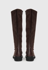 Une paire de bottes en suede brun foncé, hautes, avec des détails en cuir sur le talon et des semelles épaisses noires, vues de l'arrière sur un fond uni.