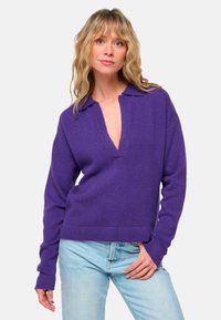 Lila gestrickter Pullover mit Kragen und tiefem V-Ausschnitt. Lange Ärmel und lockere Passform, kombiniert mit hellblauen Jeans.