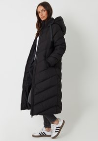 Lange zwarte pufferjas met een rits aan de voorkant, kap en diagonale stiksels. Gecombineerd met zwarte jeans en witte sneakers met zwarte strepen.
