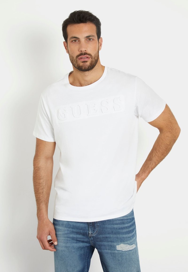 Tricou alb din bumbac cu un logo texturat "GUESS" pe față, decolteu rotund, mâneci scurte și un croi lejer, asortat cu blugi din denim.