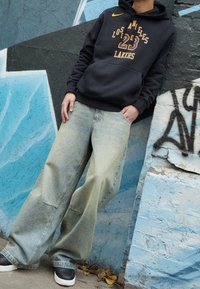 Haut à capuche noir avec "Los Angeles Lakers" et le numéro "23" en violet et jaune, associé à un jean large bleu clair et des baskets noires.