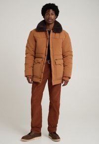 Casaco acolchoado castanho com gola de pele escura, com fecho de botões, dois bolsos frontais e punhos ajustados, combinado com calças de bombazina cor ferrugem.