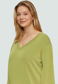 Jonge vrouw met steil blond haar, die een lichtgroene V-hals trui draagt, en zachtjes glimlacht tegen een effen lichte achtergrond.
