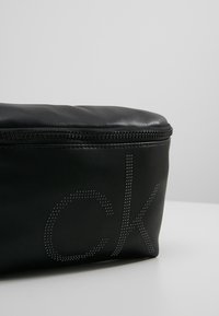 Pochette en cuir noir à la texture lisse. Dispose d'une fermeture éclair et d'un logo "ck" ponctué sur le devant. Forme rectangulaire compacte.