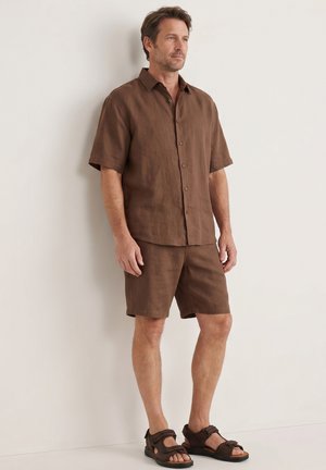 Man staat tegen een witte muur, draagt een bruin kort mouwen overhemd met knopen, bijpassende korte broek en bruine sandalen.