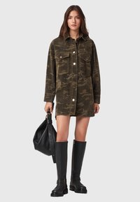 Frau trägt ein grünes Camouflage-Kordkleid, schwarze kniehohe Stiefel, hält eine große schwarze Handtasche mit dickem Trageriemen.