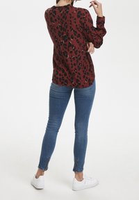 Rote Bluse mit langen Ärmeln und schwarzen Leopardenflecken, kombiniert mit blauen Skinny-Jeans mit Reißverschlüssen an den Knöcheln und weißen Turnschuhen.