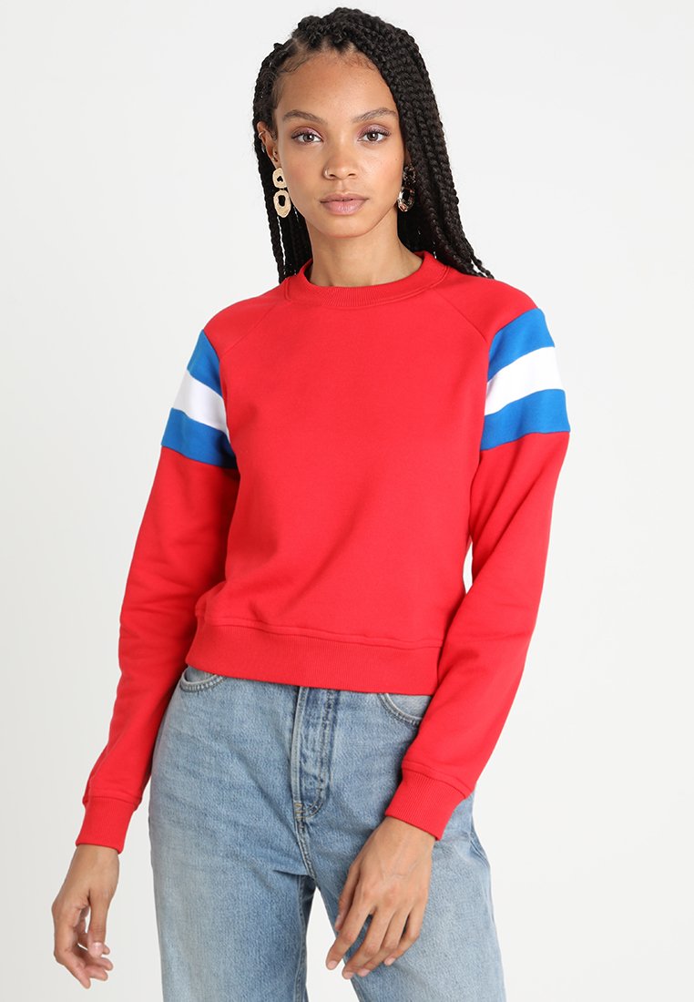 Felpa rossa con strisce blu e bianche sulle maniche, colletto e orlo a coste, realizzata in un tessuto liscio. Indossata con jeans azzurri chiari.