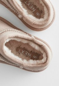 Des chaussons en tissu marron avec une doublure douce et pelucheuse, des bords arrondis et un design cousu. L'intérieur présente une étiquette avec "NEXT" en blanc.